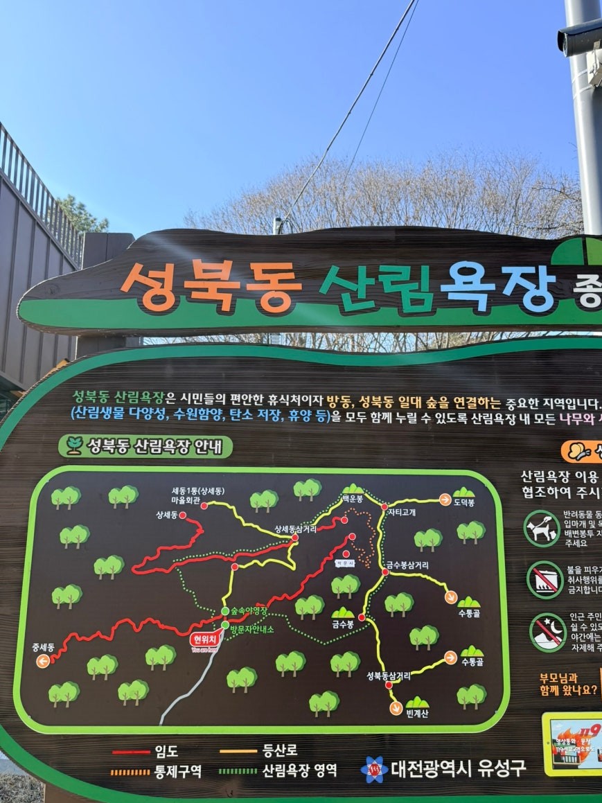 Trailhead information board at Seongbuk-dong Forest Bathing Area showing a color-coded trail map with red, yellow, and green route markings through the hills of Yuseong-gu, Daejeon, with Korean visitor guidelines listed on the right panel