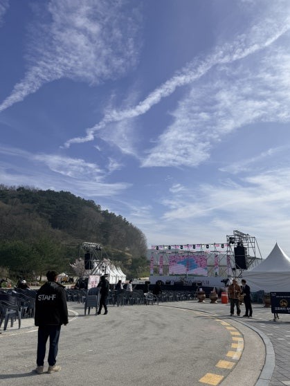Open plaza near Bungeo Island entrance with a large festival stage under construction and white tents, a staff member visible in the foreground, clear blue sky with wispy clouds above
