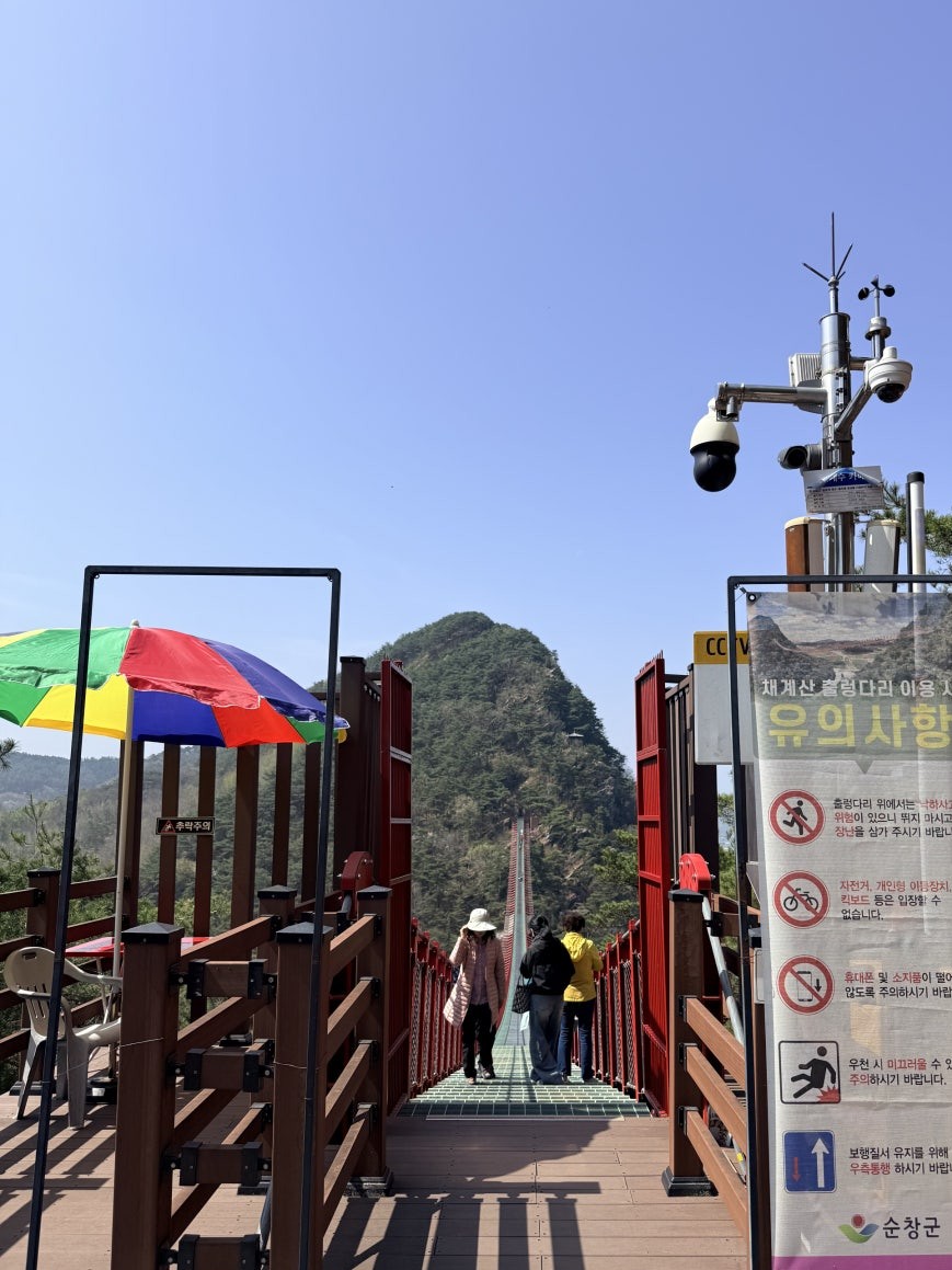 Chaegyesan Suspension Bridge entrance with red wooden railings, green metal grate walkway, Korean safety rules sign on the right, colorful umbrella on the left, two visitors stepping onto the bridge with a pointed mountain peak ahead