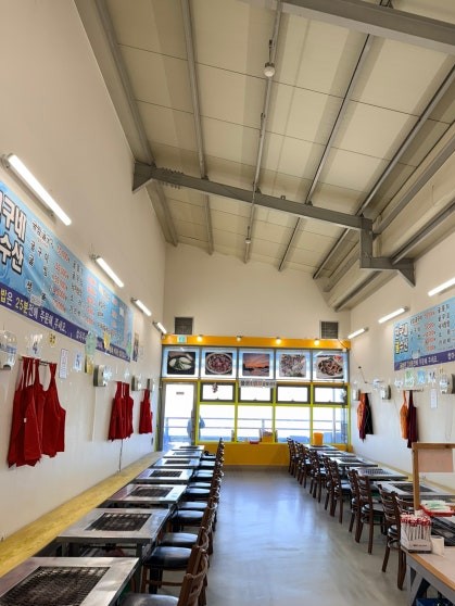 Interior of Chinguné Gul Susan with long parallel rows of tables, red aprons hanging on both walls, exposed steel ceiling beams, and menu boards visible at the far end