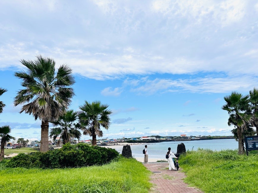 Brick-paved path through lush green grass with tall palm trees on both sides, two visitors walking toward turquoise water and a sandy beach on Udo Island under blue sky with scattered clouds