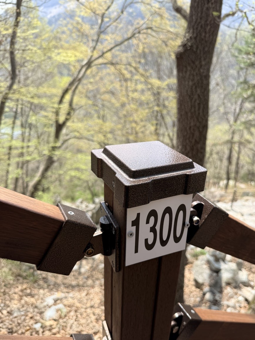Close-up of a label reading 1300 mounted on a wooden deck stair railing post, with early spring trees and a faint glimpse of the river valley in the background
