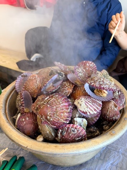 Large stone bowl heaped with steaming assorted shellfish — scallops with fan-shaped purple and pink ridged shells mixed with irregular oyster shells — steam rising above the table