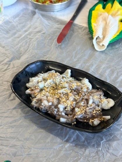 Black rectangular plate of san-nakji (산낙지, seasoned live octopus) with sliced tentacles dressed in sesame seeds, served as an appetizer on a foil-covered table