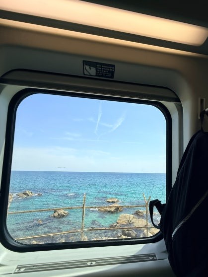 East Sea through a KTX-Eum window: clear turquoise water, a rocky shoreline with a low wooden fence, and open blue sky