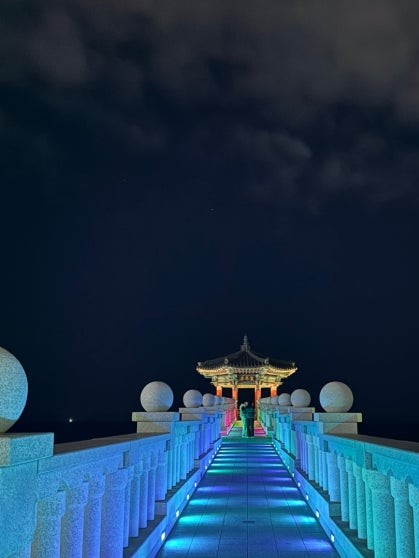 Long stone walkway with blue LED-lit pillars lining both sides, leading toward the illuminated Yeonggeumjeong sea pavilion glowing at the far end under a cloudy night sky