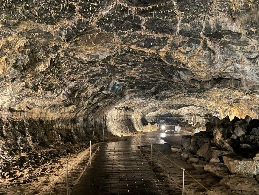 Interior of Hyeopjae Cave with a long lit brick walkway disappearing into darkness, stalactites hanging from the rough volcanic ceiling, and uneven lava rock walls on both sides