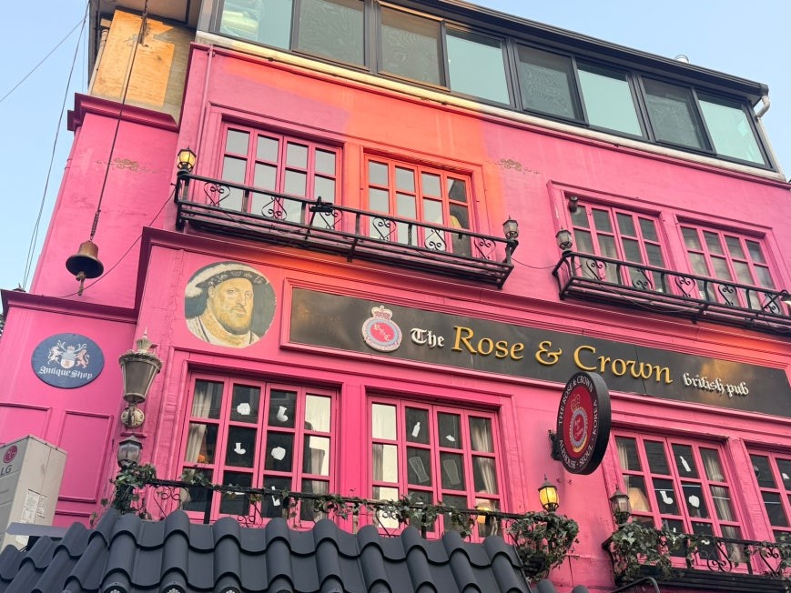 Bright pink three-story exterior of The Rose & Crown British Pub in Itaewon, Seoul, with Tudor-era portrait panels flanking the signage, wrought iron balconies, and pink-framed windows