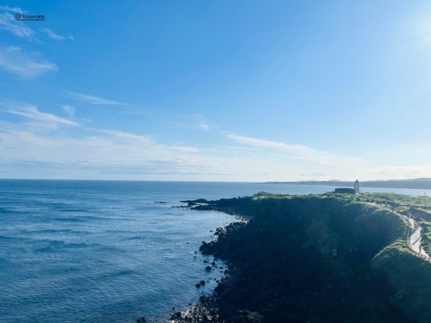 Seopjikoji (섭지코지) headland viewed from elevation — a narrow path follows the grassy clifftop toward a small white lighthouse structure, deep blue sea to the left and open coastline beyond under afternoon sun