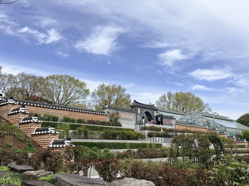 Terraced garden complex at Jeonju Arboretum with layered traditional Korean curved roof tile walls in terracotta and dark gray, a glass greenhouse running alongside the upper terrace, tiered stone steps with rock and shrub plantings, and visitors visible near a roofed arch