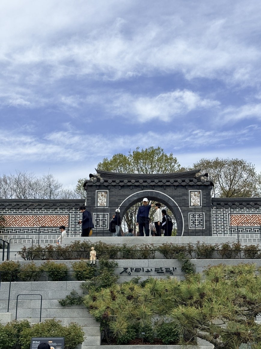 Traditional Korean circular moon gate in a dark gray brick wall with terracotta lattice tile bands and floral inlay panels at Jeonju Arboretum, visitors on top of the wall, 장미와 단림 carved in stone below, gnarled pine tree in the foreground