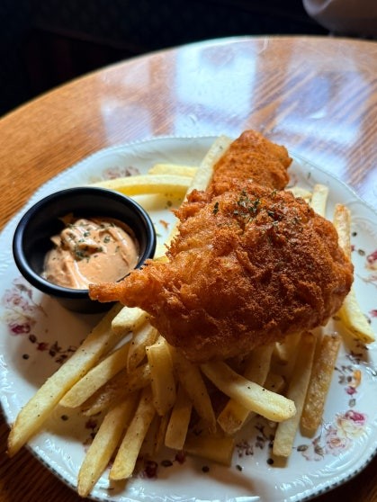 Fish and chips on a floral-patterned plate with a large golden-battered fish fillet, a pile of fries, and a bowl of orange dipping sauce at Rose & Crown