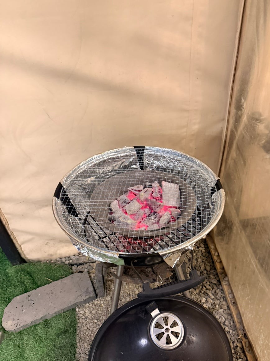 Charcoal grill at Dosim Sok Grill with glowing red embers visible through a wire mesh grate, aluminum foil lining the bowl, placed on artificial grass beside a tent canvas wall