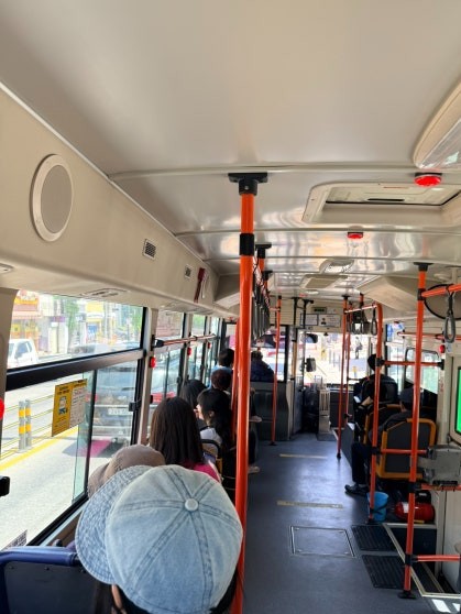 Interior of a local Mukho-area bus with passengers seated, bright orange grab poles along the aisle, and coastal scenery through the windows