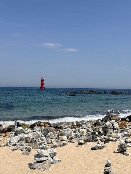 Eodal Beach: stone cairns stacked across the sandy foreground, a red offshore lighthouse buoy in the middle distance, clear turquoise East Sea water under a cloudless blue sky