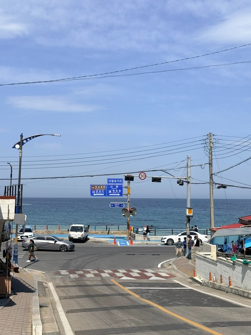 Eodal Intersection road junction with overhead blue directional signs including City Hall (시청) and Mukho Harbor, the East Sea visible straight ahead at the end of the road, parked cars on the right