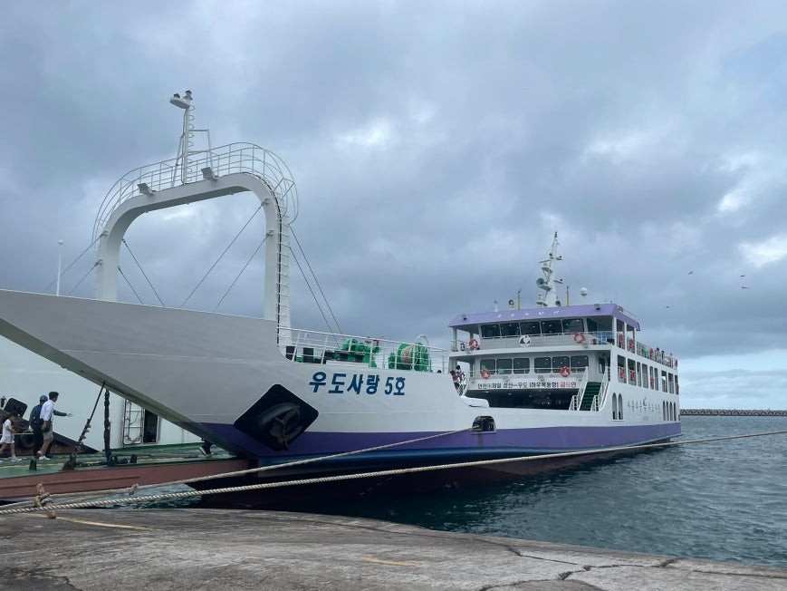 White and purple ferry named 우도사랑 5호 docked at Seongsan Port with passengers walking up the boarding gangway, calm harbor water and overcast sky in the background