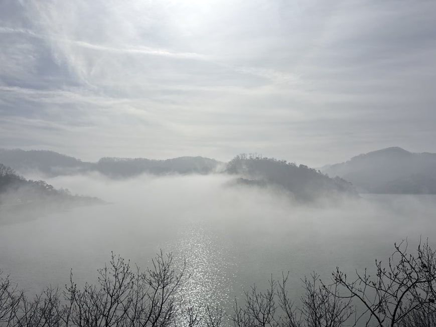 Morning mist rising over Okjeongho Lake with forested mountain ridges fading into low cloud, sunlight catching the water surface, bare winter trees in the foreground
