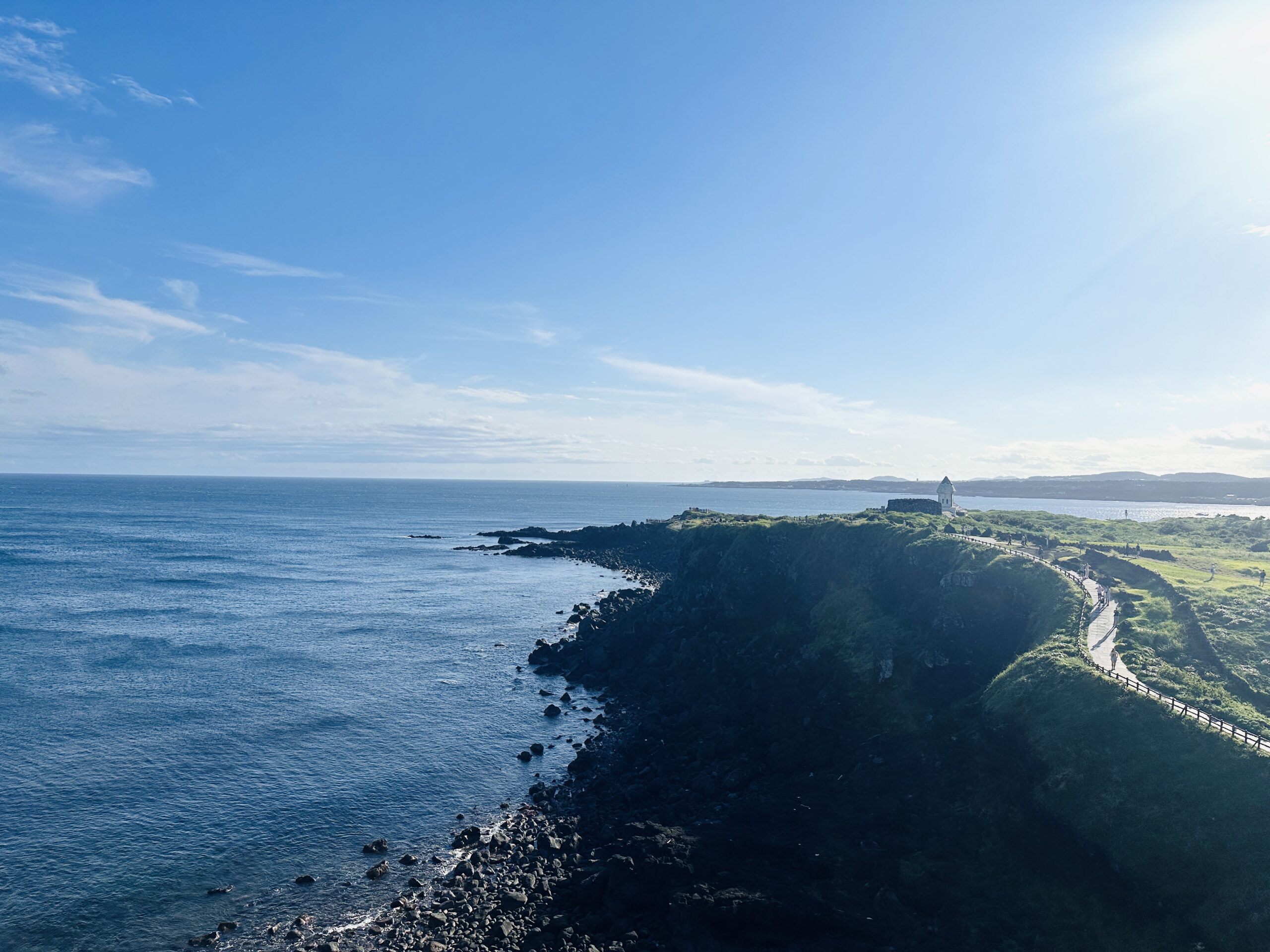 Elevated view of a green coastal headland on Jeju's south coast curving toward a small white lighthouse-like structure at the point, a narrow walking path with wooden railings following the cliff edge above dark rocks and calm blue sea