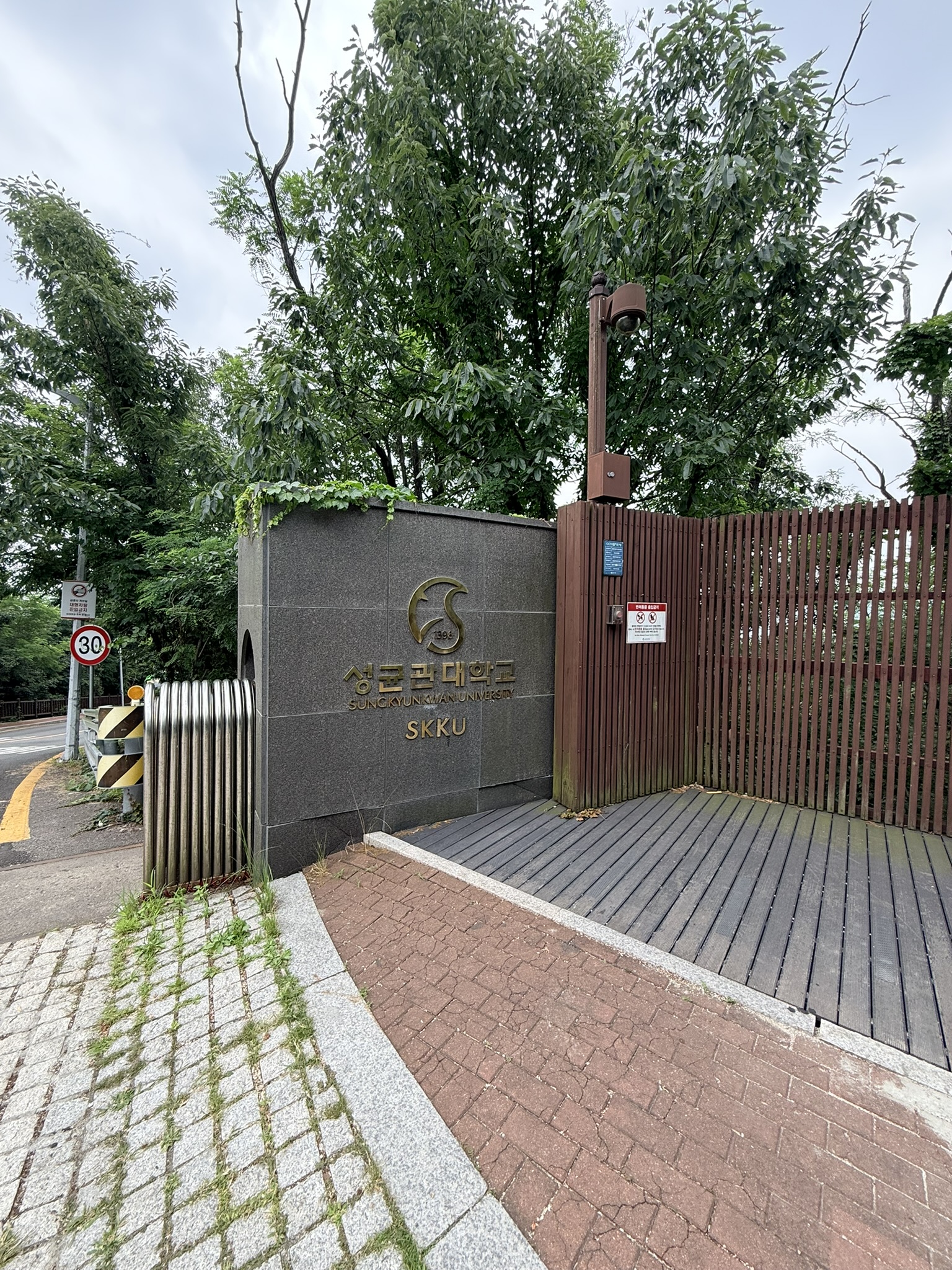 SKKU Sungkyunkwan University side gate with dark granite monolith bearing a gold university crest and SKKU lettering, brown wooden fence alongside, lush green trees overhead, on the approach road to the Bugaksan Malbawi trailhead