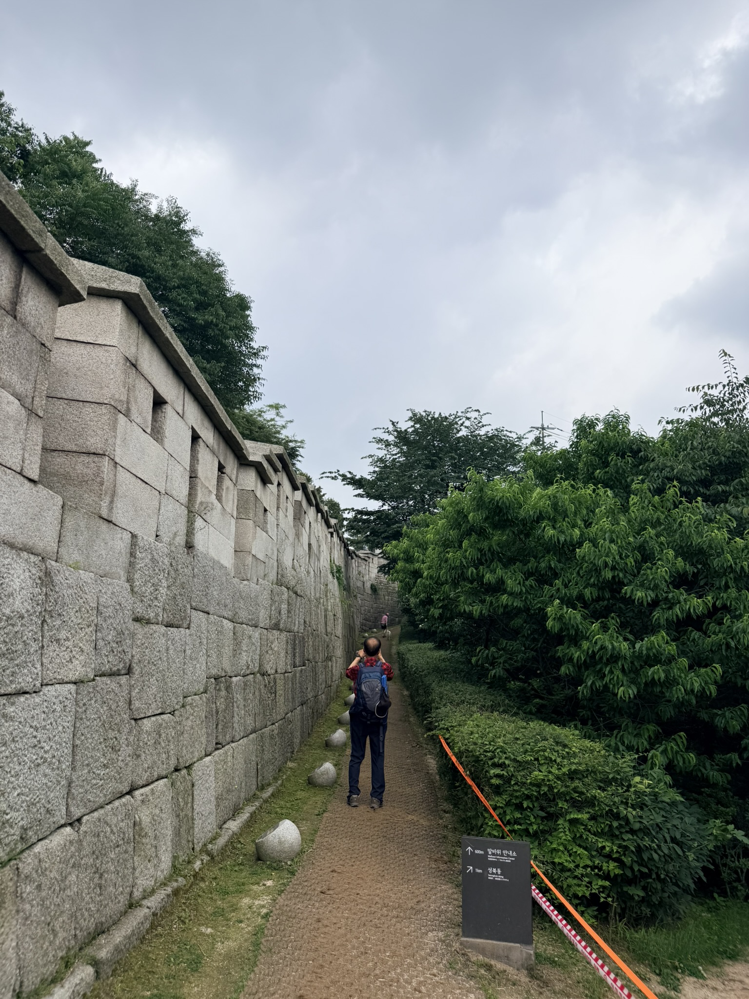 Hiker with backpack walking alongside the massive granite Hanyangdoseong city wall on the Bugaksan trail, with a dark bilingual directional signboard pointing toward Malbawi Visitor Center and Seongbuk-dong