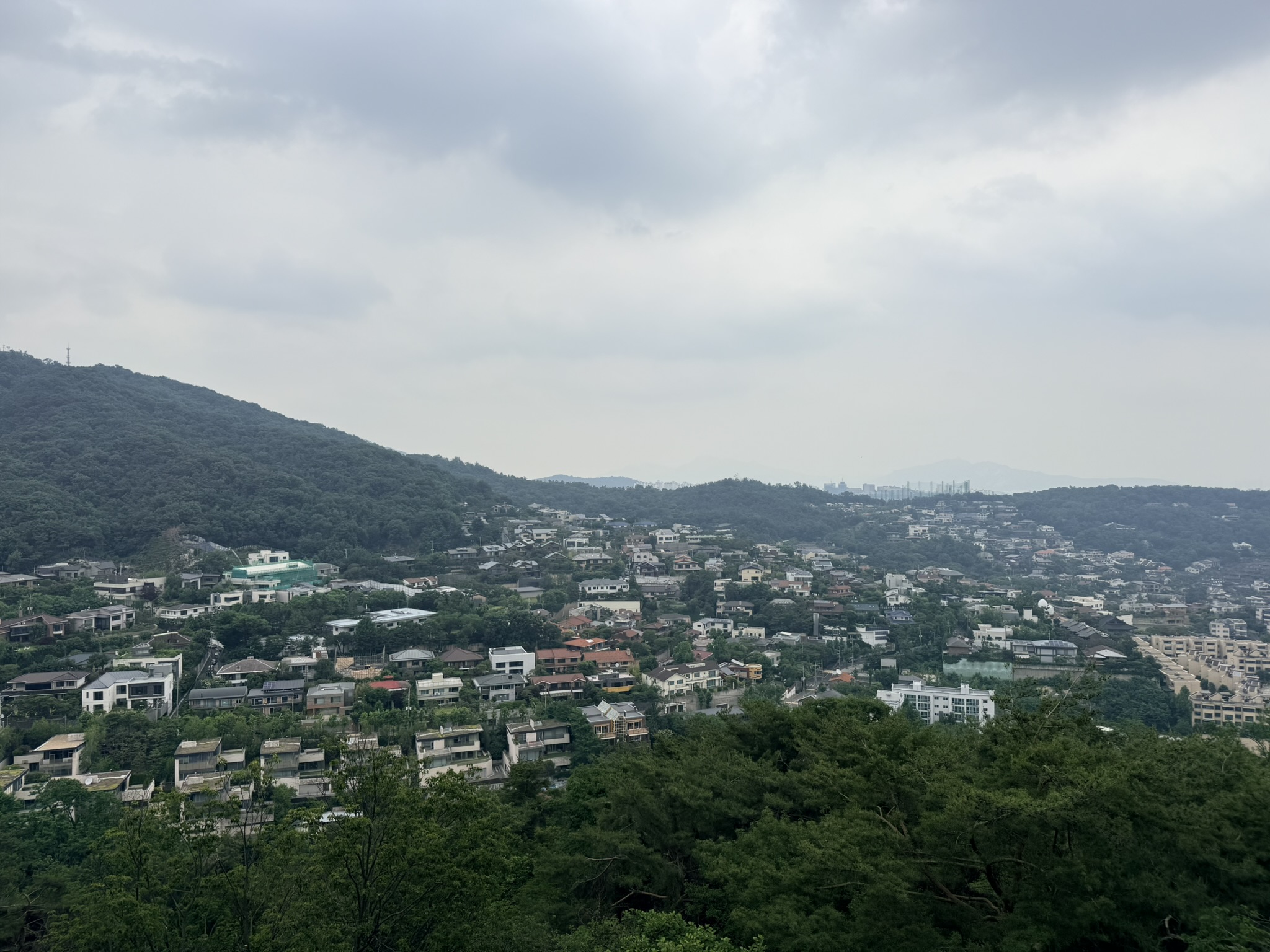 Aerial view of Seongbuk-dong hillside residential neighborhood from the Bugaksan hiking trail, densely packed houses surrounded by forested hills, distant apartment buildings on the horizon under heavy overcast sky