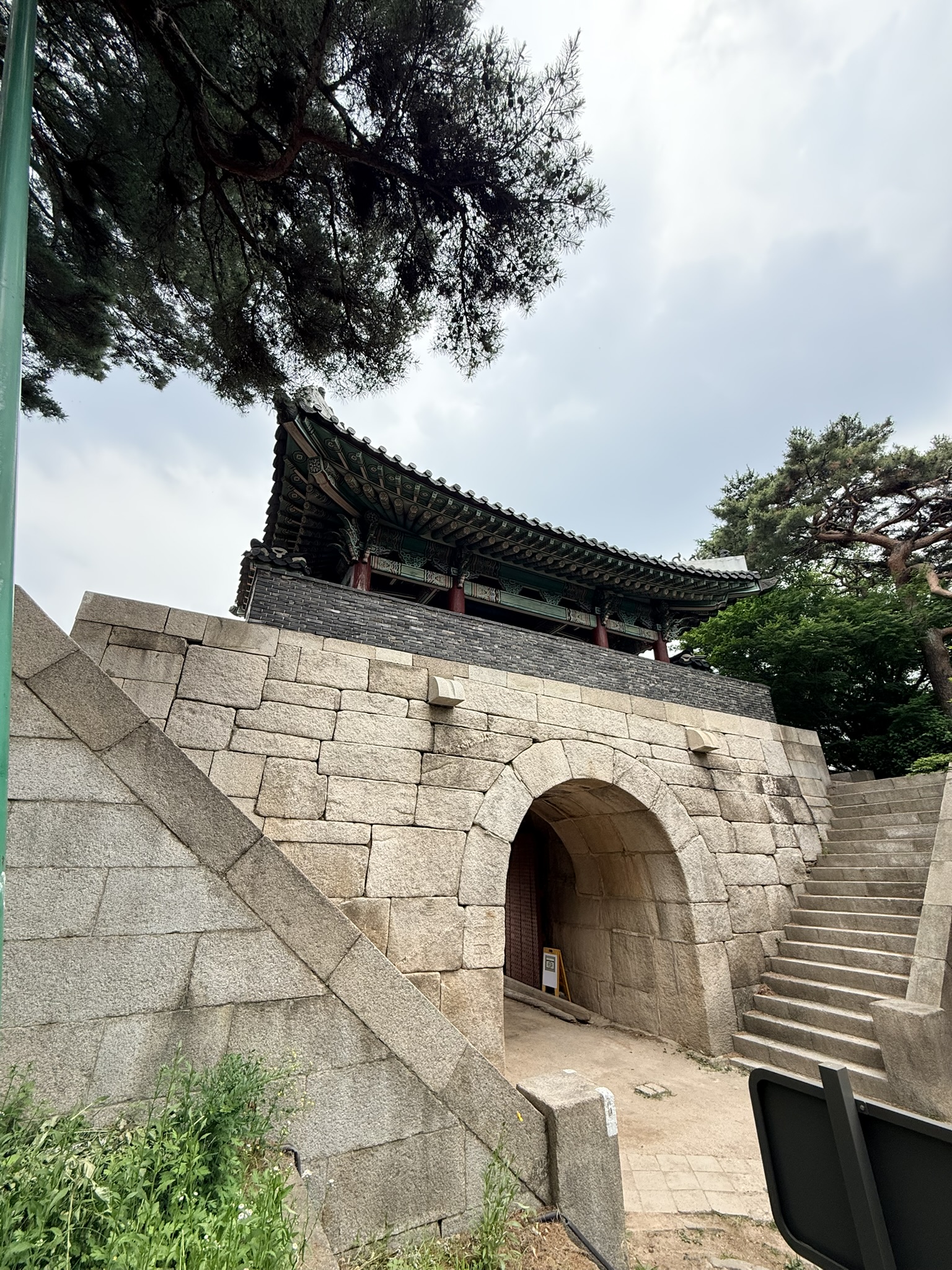 Sukjeongmun North Gate of the Hanyangdoseong city wall with rounded granite arch opening at the base, traditional wooden gate pavilion with green and black dancheong painted eaves above, stone stairs on the right, flanked by pine trees