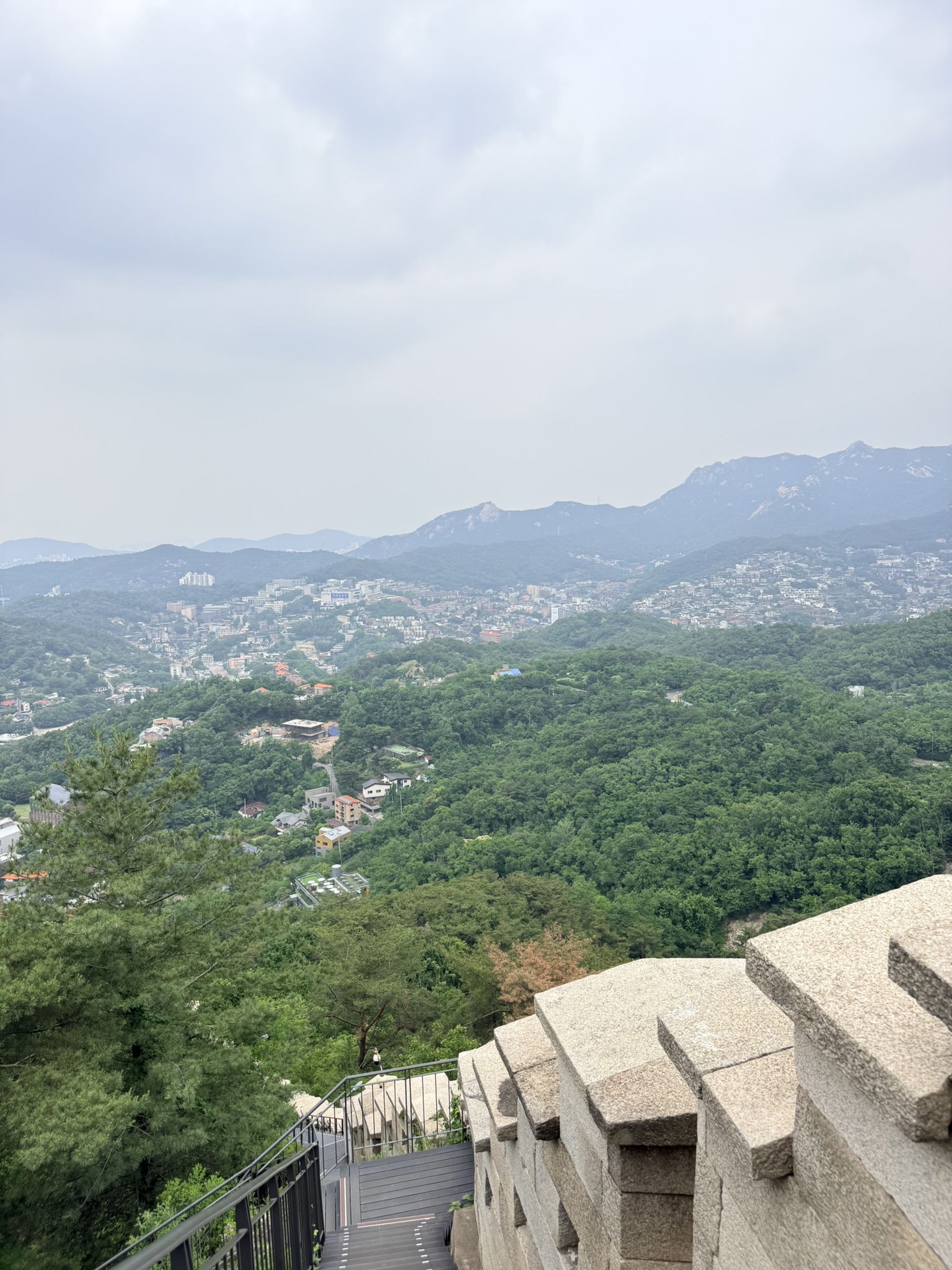 Panoramic view from near Baegaksan summit looking northwest over northern Seoul residential neighborhoods and the Bukhansan mountain range, granite city wall crenellated battlements in the foreground and dense forest in the middle ground