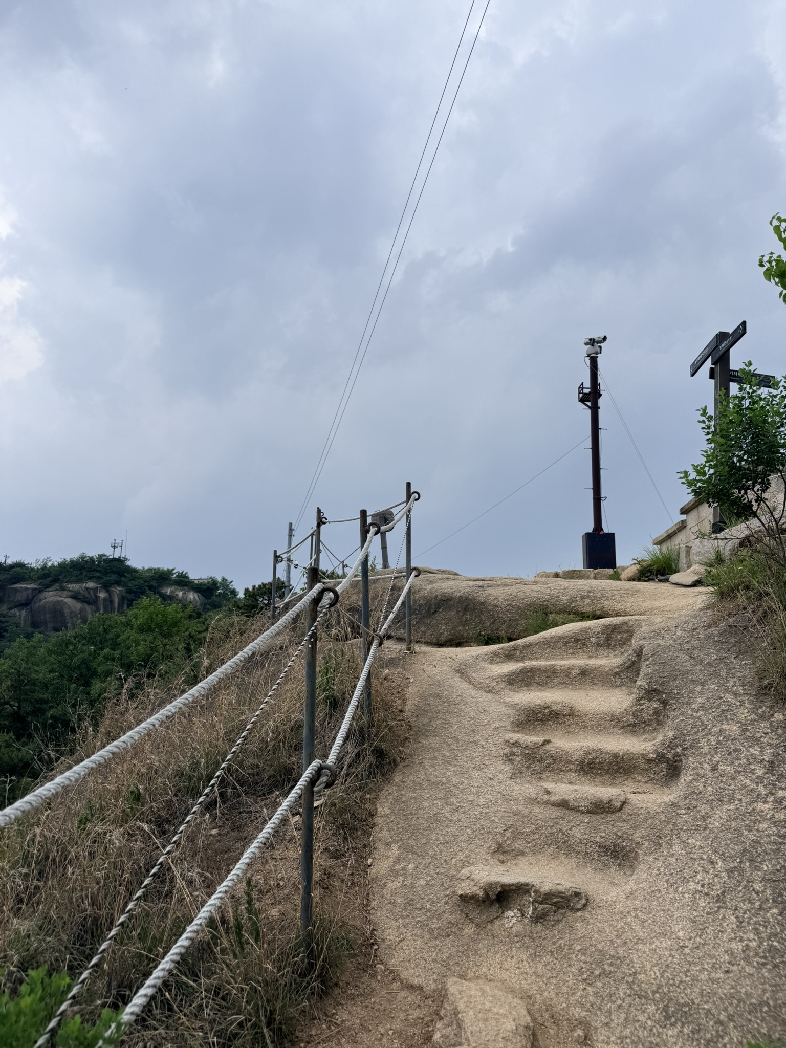Steps carved into natural granite with rope railings on the upper Inwangsan trail, dark storm clouds building overhead, CCTV pole and trail signpost visible at the top right