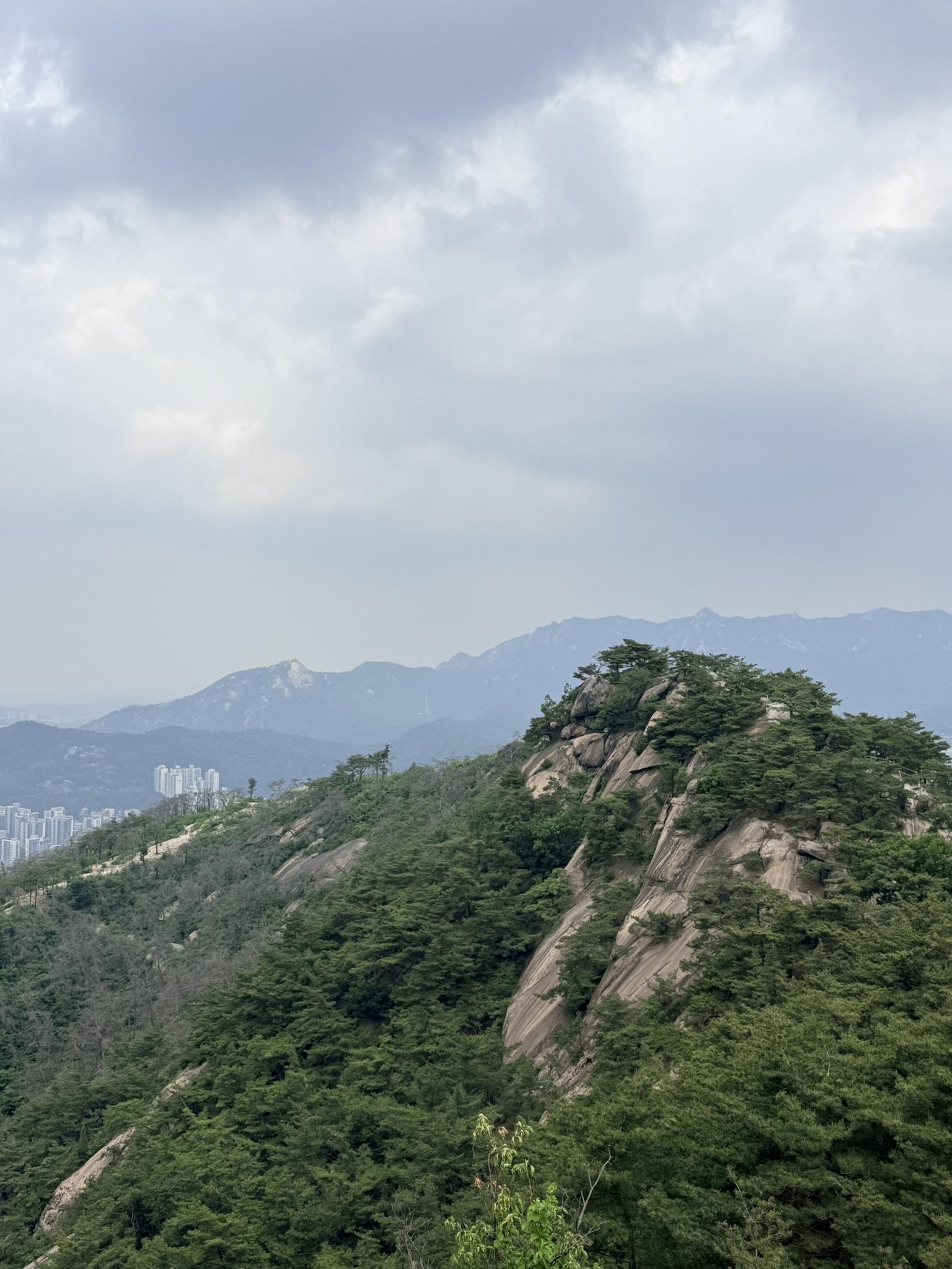 Exposed granite ridge and dome of Inwangsan with pine trees clinging to the rock surfaces, layered mountain ranges and high-rise buildings visible through overcast haze in the background