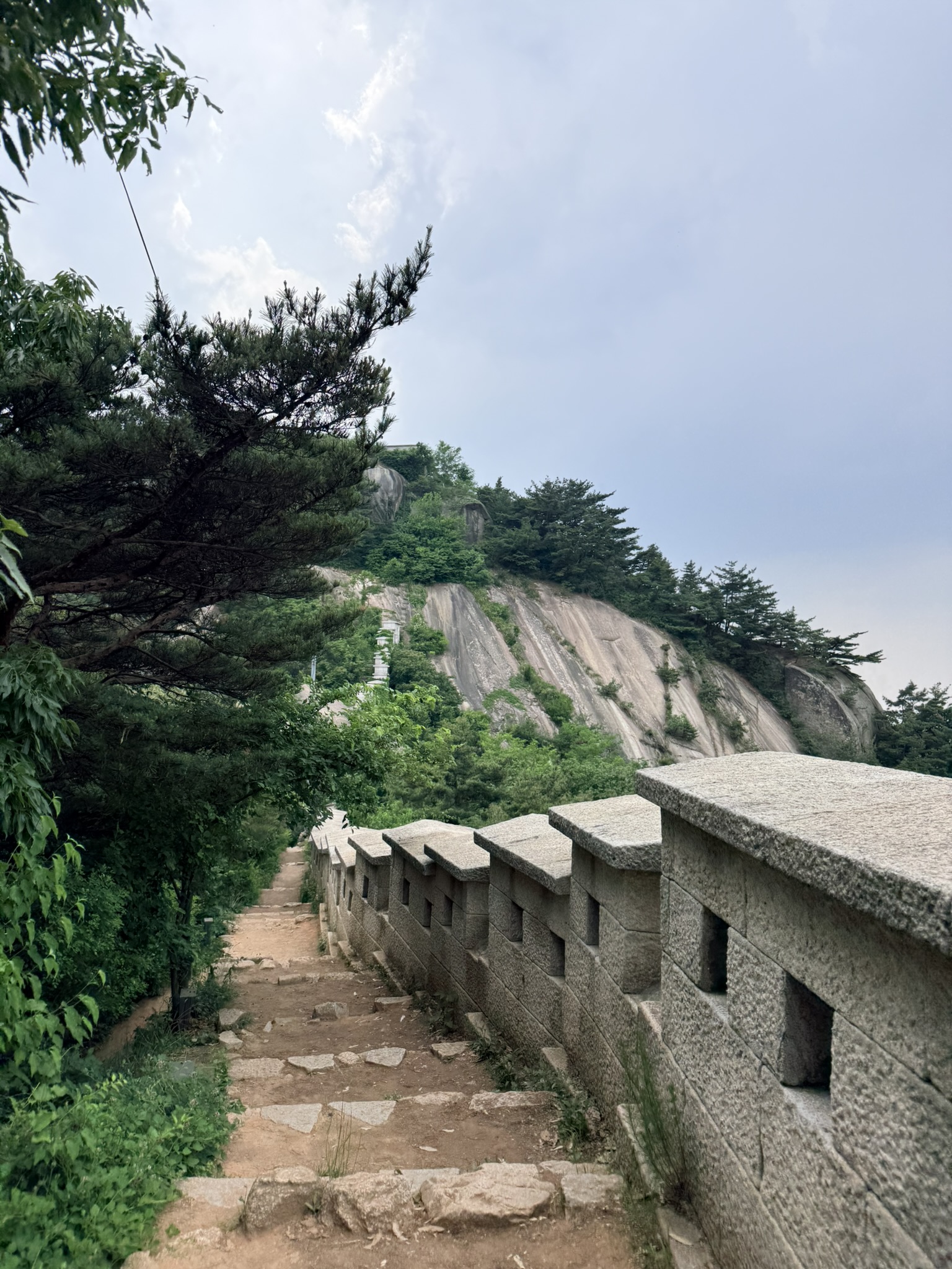 Dirt and stone staircase running alongside Hanyangdoseong battlements with rectangular embrasures, large granite dome and pine trees visible above on Inwangsan