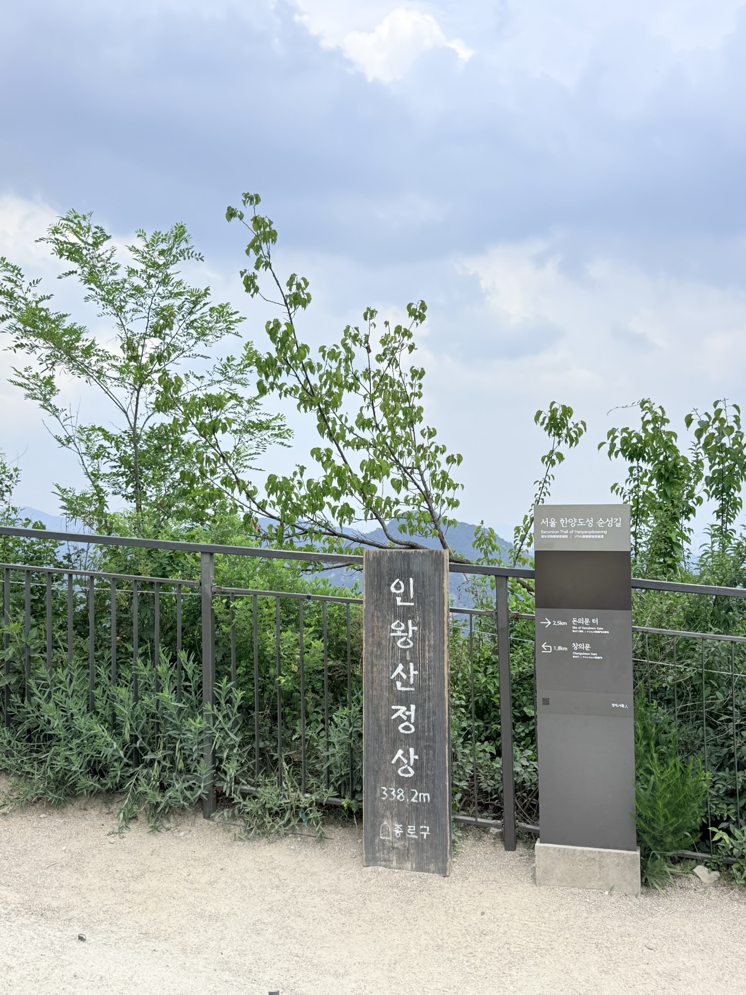 Inwangsan summit marker 인왕산정상 338.2m alongside Seoul Hanyangdoseong trail sign showing directions to Donuimun Gate site 2.5km and Changuimun Gate 1.8km, leafy vegetation in the background