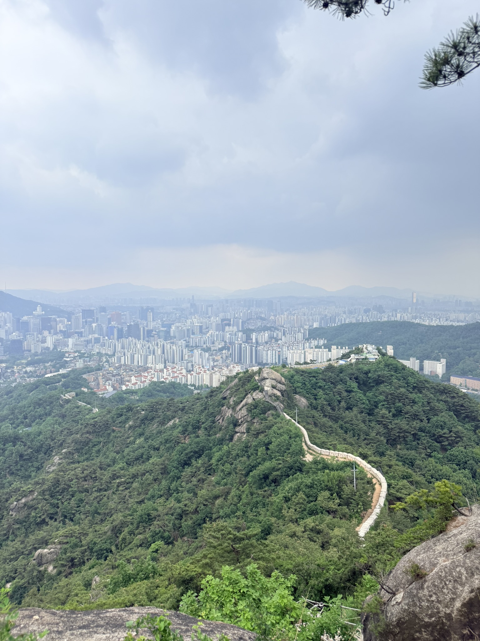View from Inwangsan summit looking south along Hanyangdoseong city wall snaking along the ridge below, Seoul cityscape filling the valley under overcast sky, pine branch framing the upper right corner